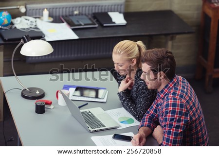 Motivated team at work in the office with a young businessman and woman sitting at a desk smiling as they read information on a laptop together, high angle view