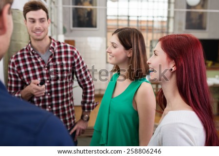 Group of young people or co-workers standing together chatting and laughing in the office with focus to a smiling young man at the rear of the team