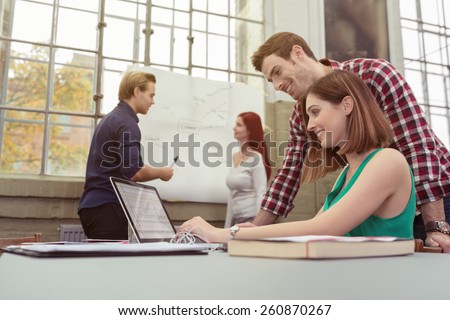 Two business colleagues in a busy modern office with motivated young people working together on a laptop computer smiling as they read information on the screen