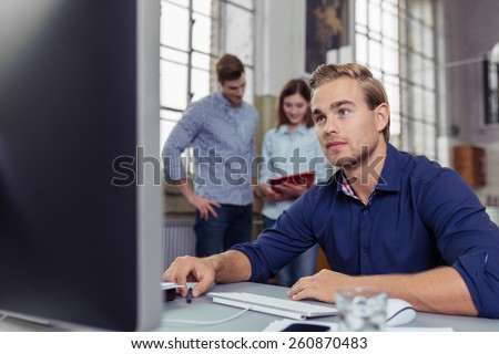 Busy office with a hardworking young man sitting at his desktop computer in the foreground with his colleagues have a discussion over paperwork in the background