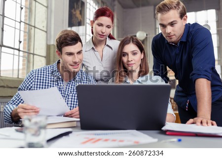 Young business team analysing company data sitting and standing together in a group at a desk with a computer and paperwork having a discussion