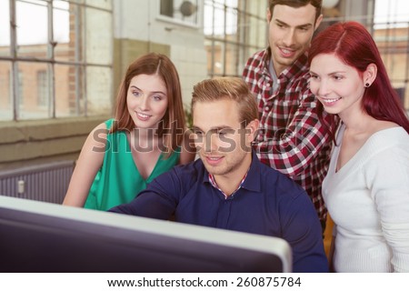 Smiling group of young people in the office clustered around a young man at a desktop computer reading the information on the screen