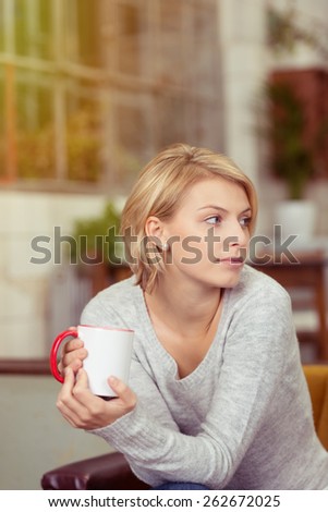 Observant young woman with a cup of coffee cradled in her hands sitting staring off to the right of the frame