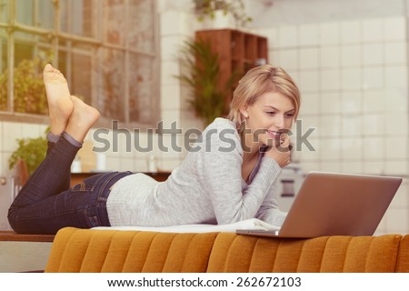 Happy Blond Woman Lying on her Stomach on the Table Behind the Couch While Watching a Movie at her Laptop Computer.