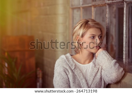 Young woman sitting daydreaming or waiting for someone staring out of a window in the living room with a serious expression