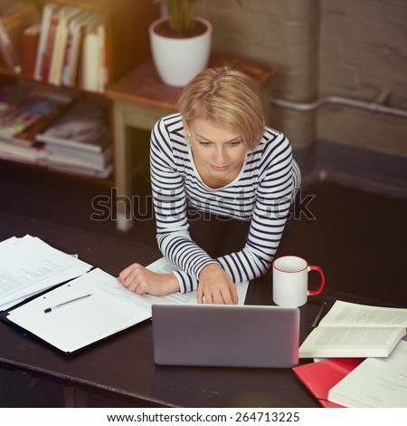 Pretty Blond Woman Leaning on the Table While Working on her Laptop with Documents, Books and Coffee Mug on the Sides.