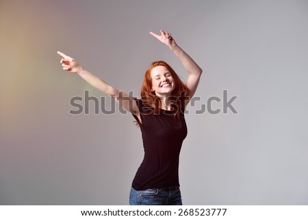 Happy Young Red Haired Woman in Motion, Wearing Casual Black Shirt and Jeans, Pointing Upper Right with Closed Eyes on a Gray Background.