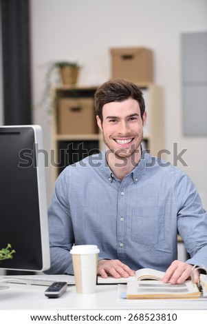 Young Handsome Businessman Sitting at his Worktable with Books, Computer and a Cup of Coffee While Smiling at the Camera
