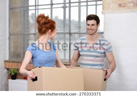 Loving couple moving house carrying cardboard boxes as they work as a team pausing to smile at one another