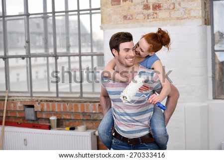 Loving young woman getting a piggy back ride from her handsome husband as she nuzzles him on the cheek while holding a paint roller indoors in the apartment