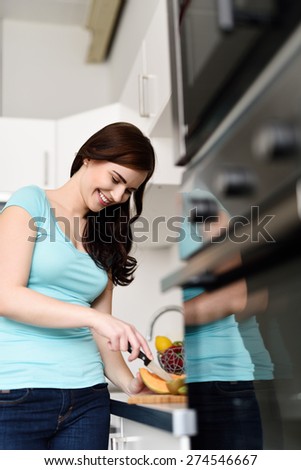 View along the cabinetry of an attractive smiling young woman chopping fruit on the counter in the kitchen