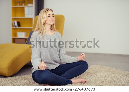 Pretty young woman practicing yoga in her living room sitting in the lotus position meditating with a serene smile and closed eyes