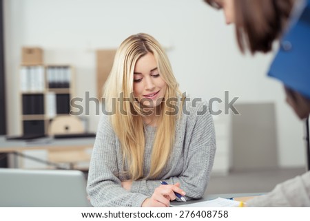 Pretty young businesswoman at work in the office sitting at her desk looking down at paperwork with a colleague with a smile