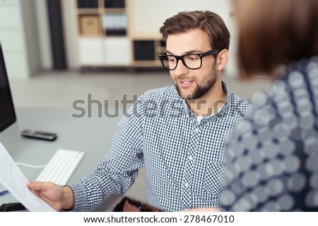 Young Businessman Wearing Eyeglasses Holding Sheet of Paper and Discussing with Co-Worker, as seen from Perspective of Behind Co-Worker