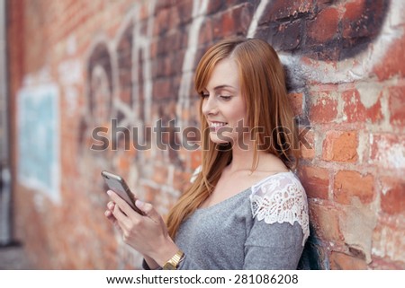 Young redhead woman smiling and leaning against a brick wall with graffiti text while looking at the touch screen of her smart phone, concept of funny networking and social media