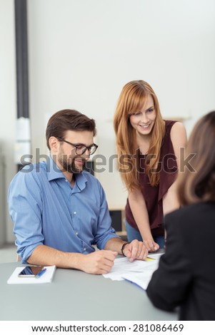 Three business people in a meeting in the office with focus to an attractive redhead woman standing leaning on the table alongside a male colleague
