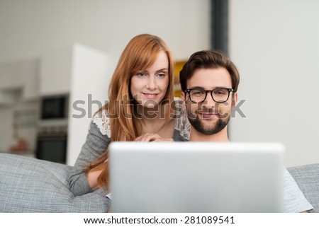 Man with Facial Hair and Glasses Sitting on Sofa Using Laptop Computer with Red Haired Woman Looking Over Shoulder at Screen Leaning Over Back of Sofa