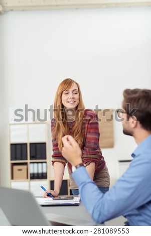 Smiling businesswoman having a discussion with a seated male co-worker as she stands leaning on a table in the office, view over his shoulder