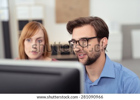 Young bearded man with black frame eyeglasses working together with his female colleague, a redhead pretty woman, in front of a desktop computer, in the office