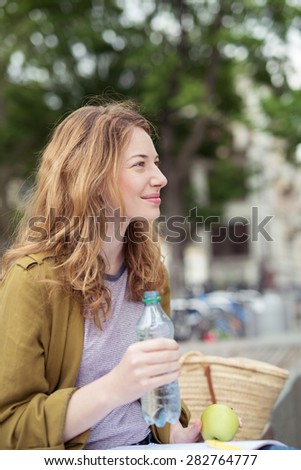 Smiling Pretty Girl Studying at the Bench, Holding Fresh Green Apple and a Bottle of Water While Looking Into the Distance.
