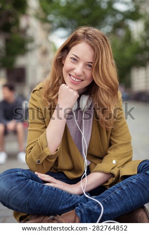 Pretty Blond Girl with Headphone Around her Neck, Sitting on Bench with Legs Crossed and Chin Resting on Hand While Smiling at the Camera.