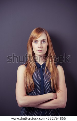 Serious Portrait of a Young Blond Woman with Arms Crossed Over her Stomach, Staring at the Camera on a Gray Background
