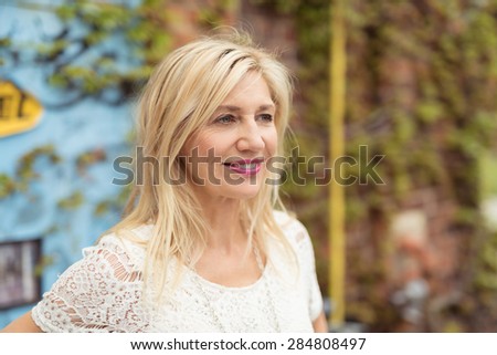 Attractive middle-aged blonde woman with medium-length hair and white laced T-shirt looking away with a serene and jovial facial expression, outdoors, in front of a wall with green climbing plants