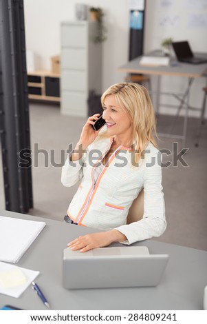 Happy Blond Adult Businesswoman, Sitting at her Table with Laptop, Talking to Someone on Mobile Phone