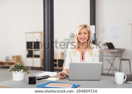 Happy Blond Adult Businesswoman Sitting at her Table with Laptop Computer and Other Office Stuffs.