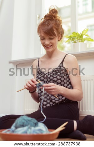 Smiling young woman sitting on the floor in front of a radiator knitting with blue wool, low angle view with focus to her happy face