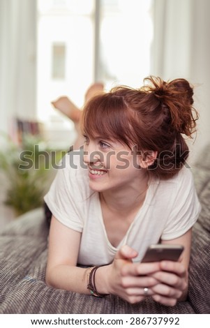 Happy young woman relaxing at home lying on the sofa with bare feet holding her mobile and looking to the side with a smile