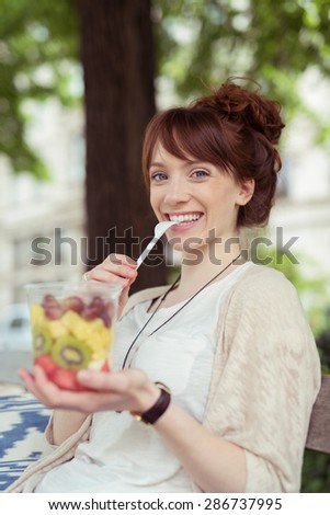 Happy woman eating fresh fruit salad with exotic tropical fruits as she relaxes the shade of a tree in a park