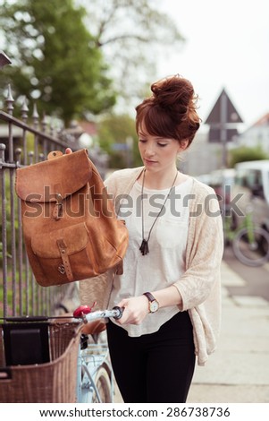 Pretty Young Woman Putting her Brown Leather Backpack on a Basket In Front her Bicycle.