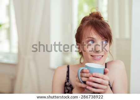 Pretty redhead woman enjoying a cup of coffee sitting clasping the mug in her hands and looking out of the window with a smile