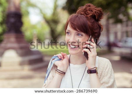 Vivacious young woman chatting on her mobile smiling with delight as she listens to the conversation outdoors in a park