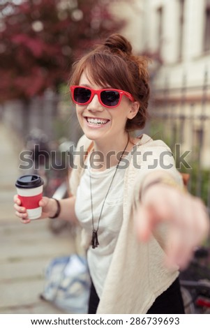 Laughing vivacious woman in chic red sunglasses pointing at the camera with a smile as she holds a cup of takeaway coffee in an urban street, focus to her face