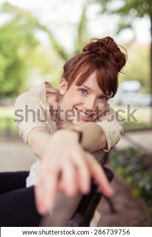 Happy smiling woman relaxing on a park bench stretching her arms along the top as she looks at the camera with a friendly smile