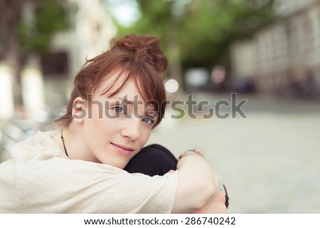 Pensive young woman gazing at the camera with a serious expression as she sits hugging her knees outdoors with copyspace