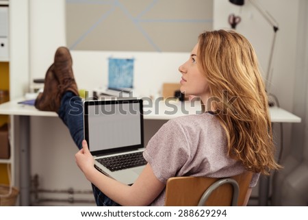 Thoughtful Blond Teen Girl Holding her Laptop Computer, Sitting at her Study Room with Feet Up on the Table and Looking Up.
