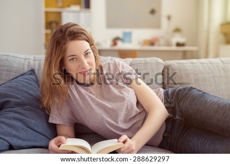 Young woman lying on a sofa with a book smiling at the camera as she spends a relaxing day at home