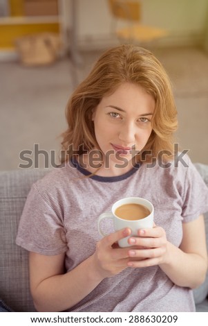 Attractive young woman sitting on a comfortable sofa clasping a mug in her hands looking up at the camera drinking delicious freshly brewed cup of coffee