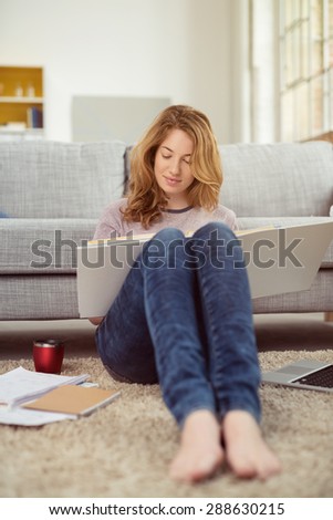 Young girl studying her class work at home as she relaxes barefoot on the living room floor with a mug of coffee, class binder and laptop