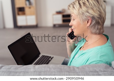 Close up Side View of a Senior Woman with Laptop Computer, Talking to Someone on Phone at the Living Room.