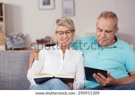 Sweet Matured Couple Sitting at the Living Room Couch with Book and Tablet Computer.