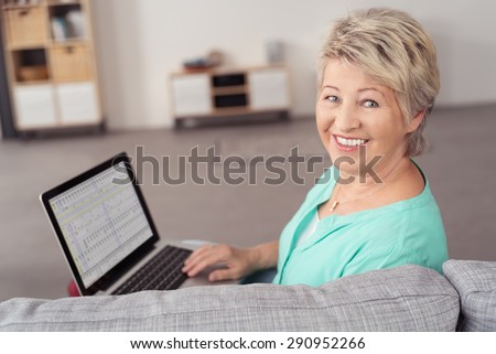 Cheerful Senior Woman Sitting at the Couch with Laptop Computer, Looking at the Camera with Toothy Smile.