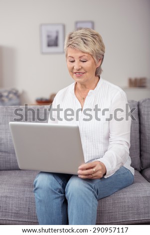 Happy Middle Aged Blond Woman Using her Laptop Computer While Sitting at the Couch Inside the House.