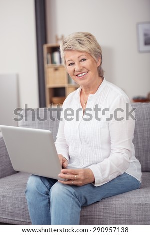 Happy Middle Aged Blond Woman Holding her Laptop Computer While Sitting on the Couch and Smiling at the Camera.