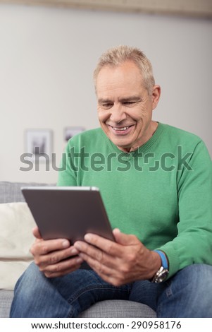 Close up Smiling Middle Aged Guy Looking at the Screen of his Tablet Computer on his Hand, While Sitting at the Couch in the Living Area.