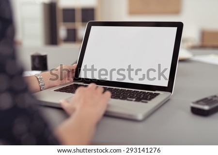 Businesswoman sitting at her desk navigating the internet on a laptop computer using the trackpad, over the shoulder view of the blank screen