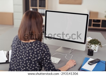 Businesswoman working on a large screen desktop computer while sitting at her desk at the office, view from behind sowing the blank monitor